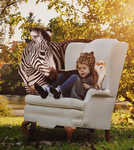 A young child is reading a book on a white chair with a zebra and owl next to her in nature for an education or creativity concept.